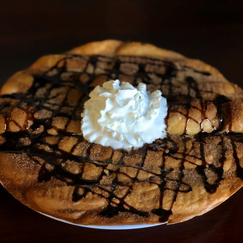A Mexican dessert, likely a sopapilla, topped with whipped cream, chocolate syrup, and cinnamon sugar.