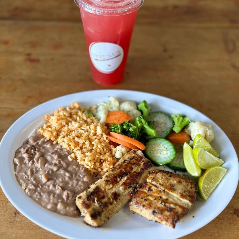 Grilled chicken with rice, refried beans, mixed vegetables, lime wedges, and a pink drink.