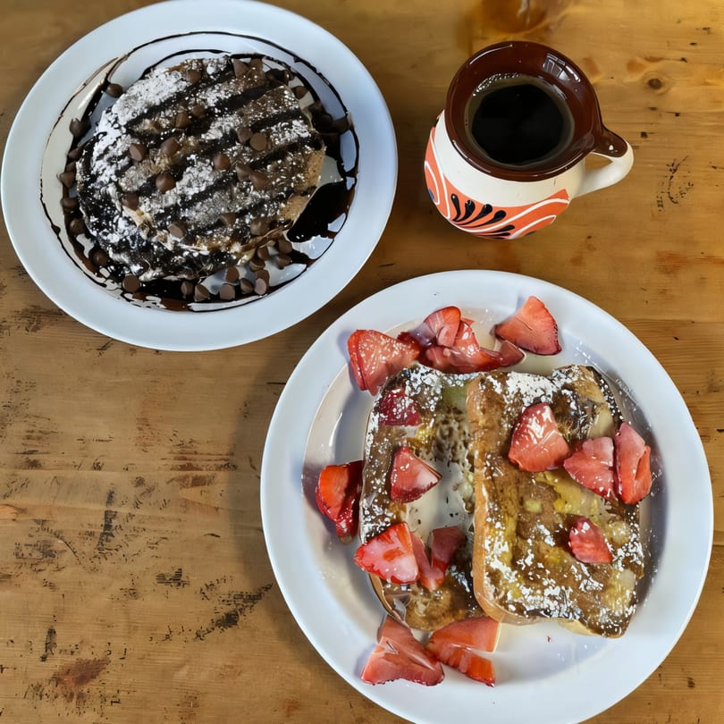 Chocolate chip pancakes, strawberry French toast, and a cup of coffee on a wooden table.