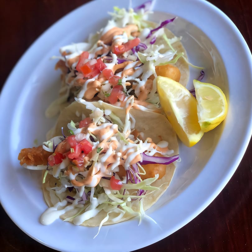 Two fish tacos with shredded cabbage, diced tomatoes, and creamy sauces, served with lemon wedges.