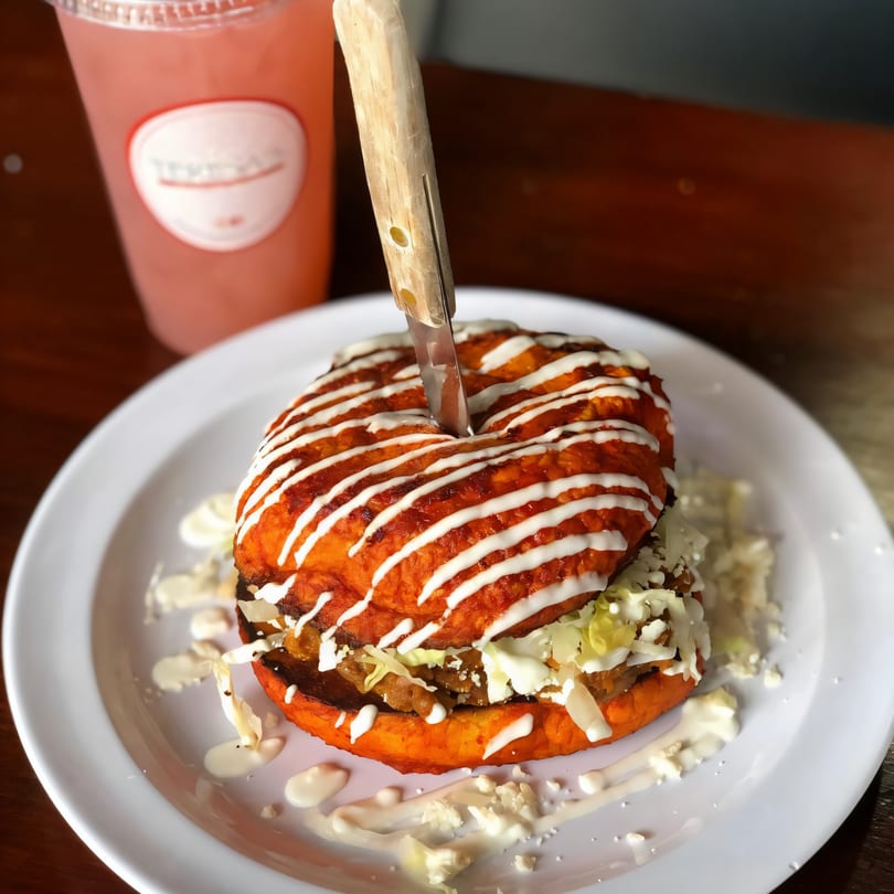 A vibrant Mexican torta with a knife, drizzled with crema, alongside a pink drink.